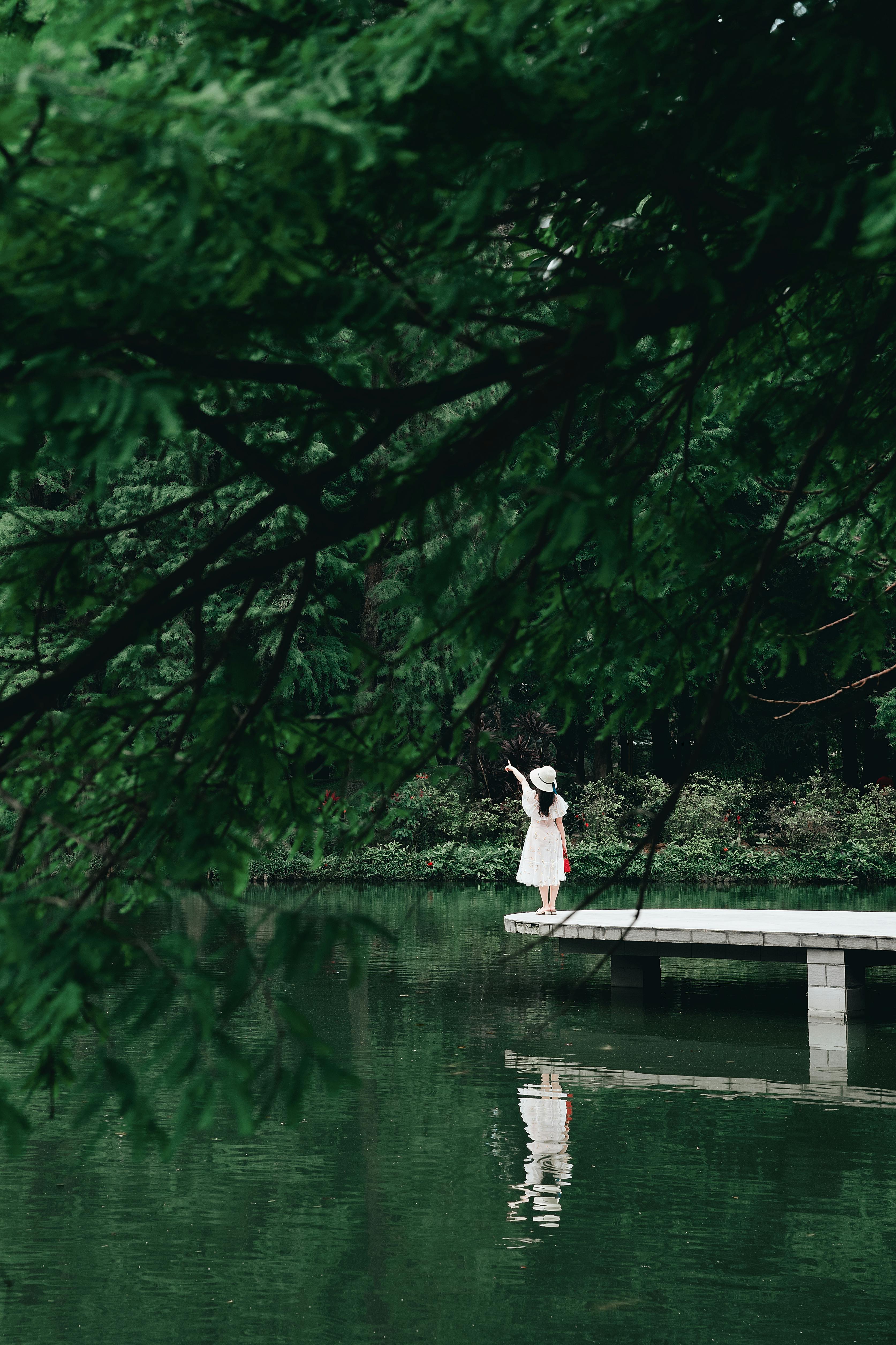 A woman in a white dress stands on a jetty, pointing amidst lush greenery by a tranquil lake.