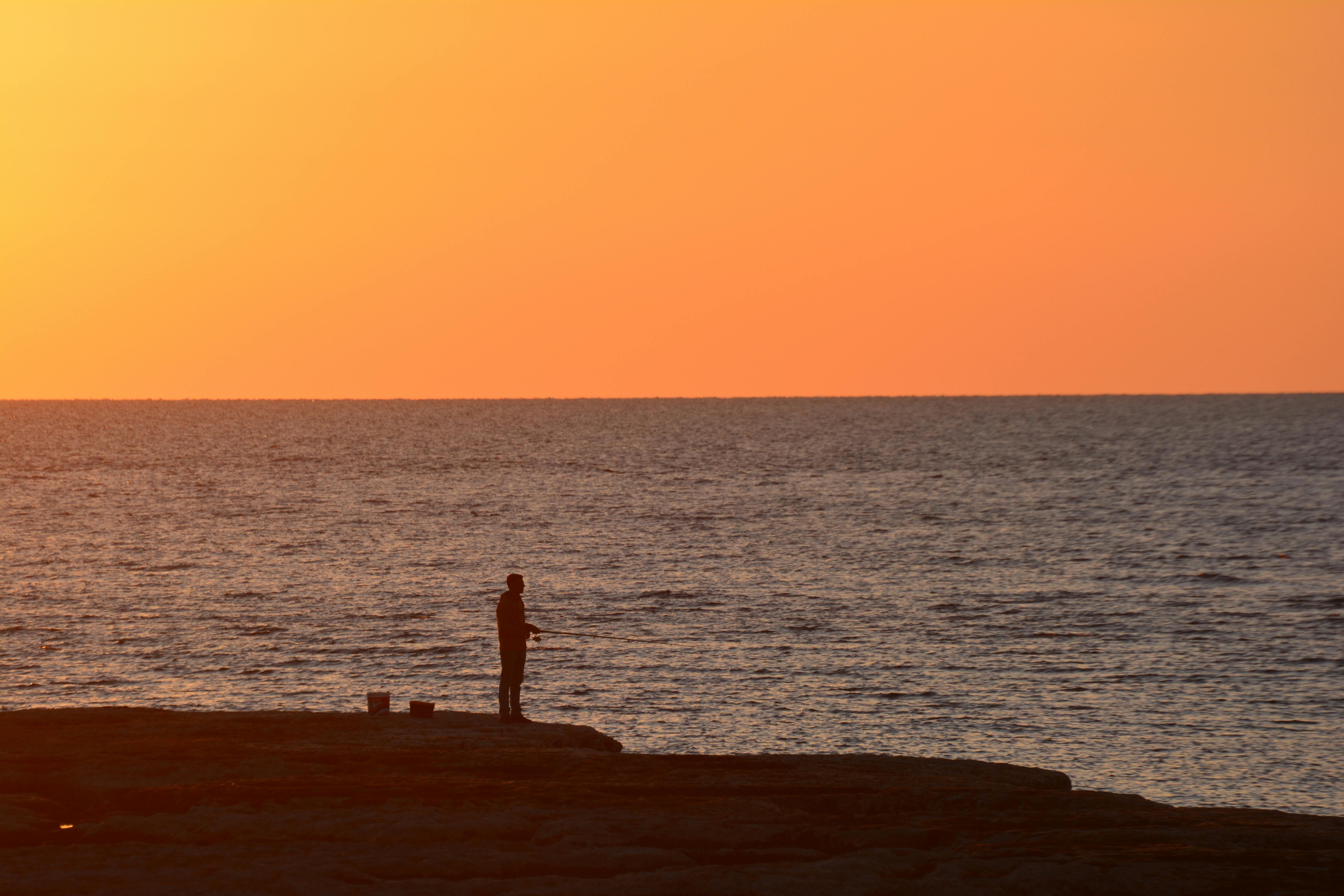 Silhouette of a fisherman by Byblos coast with a vibrant sunset sky.