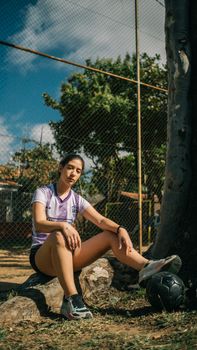 A woman in a sports jersey sits against a tree with a football, enjoying a sunny day outdoors.