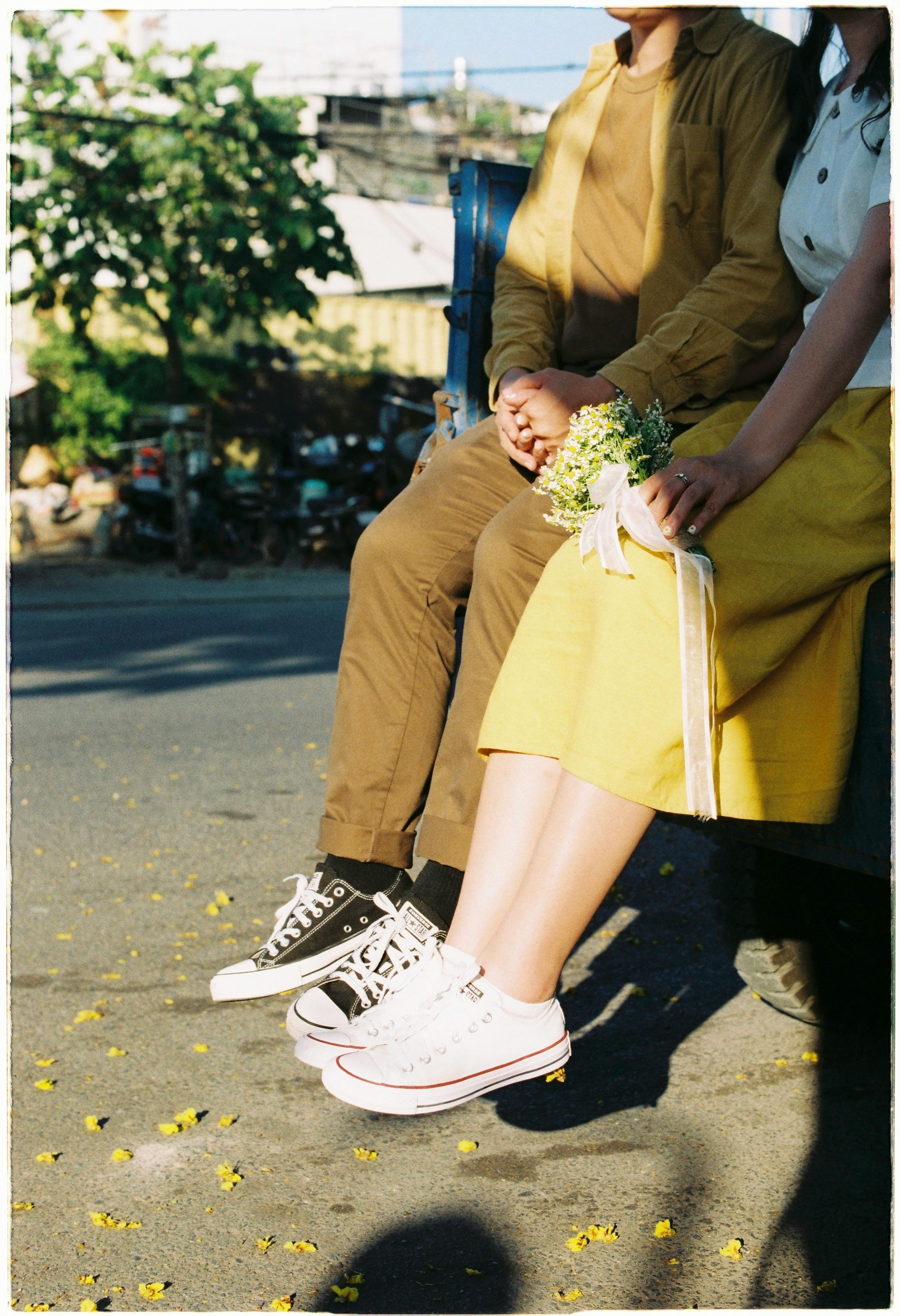 A couple sitting on a ledge outdoors, holding a bouquet with legs dangling in sunlight.