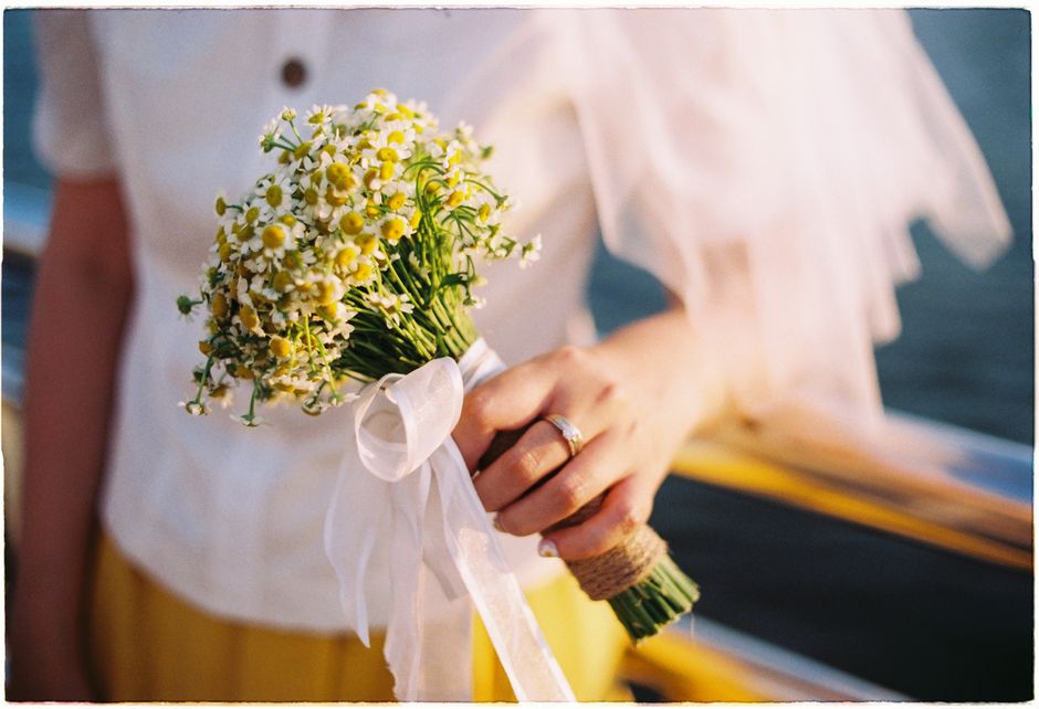 Close-up of bride's hand holding a floral bouquet with a wedding ring visible, outdoors.