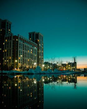 Vivid evening cityscape with buildings reflecting in water, against a dramatic night sky.