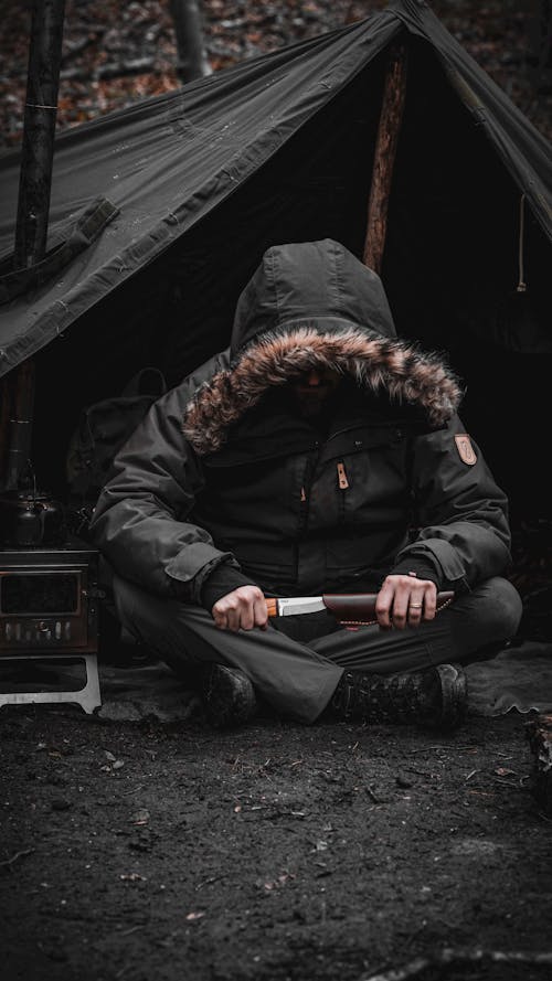 Man in a Jacket Sitting in front of a Tent and Holding a Knife 