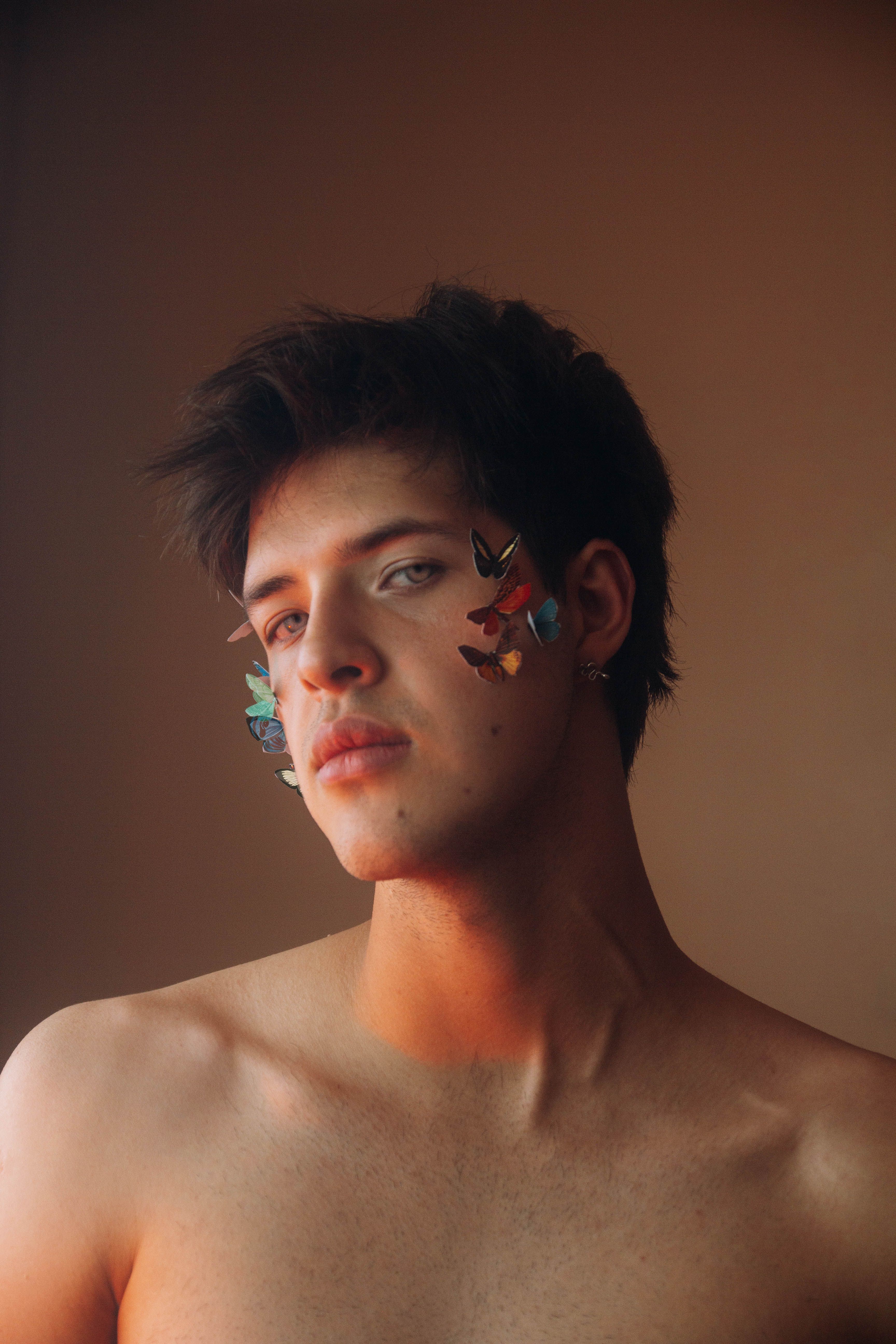 Creative portrait of a topless man with decorative butterflies on his face against a warm brown background.