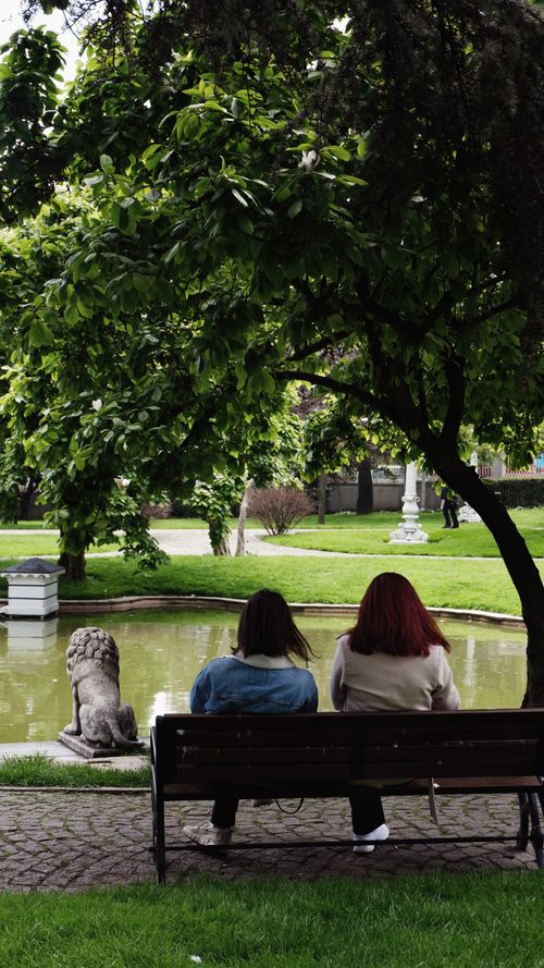 Free Two women sit on a bench, enjoying a peaceful urban park setting by a pond. Stock Photo