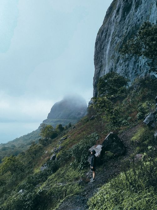 Free A hiker stands on a misty mountain trail beside towering cliffs, embracing nature's beauty. Stock Photo