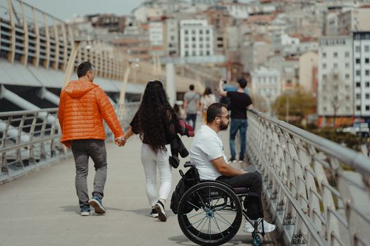 A couple and a man in a wheelchair enjoying a walk on a city bridge with urban backdrop.