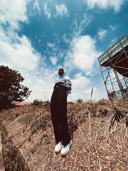 A woman in fashionable attire poses confidently on a steep roadside hill under a bright summer sky.