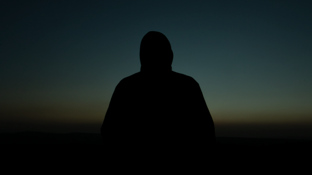 Silhouette of a hooded man standing outdoors at sunset, creating a mysterious vibe.