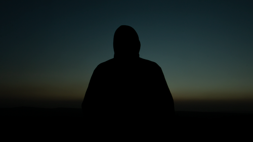 Free Silhouette of a hooded man standing outdoors at sunset, creating a mysterious vibe. Stock Photo