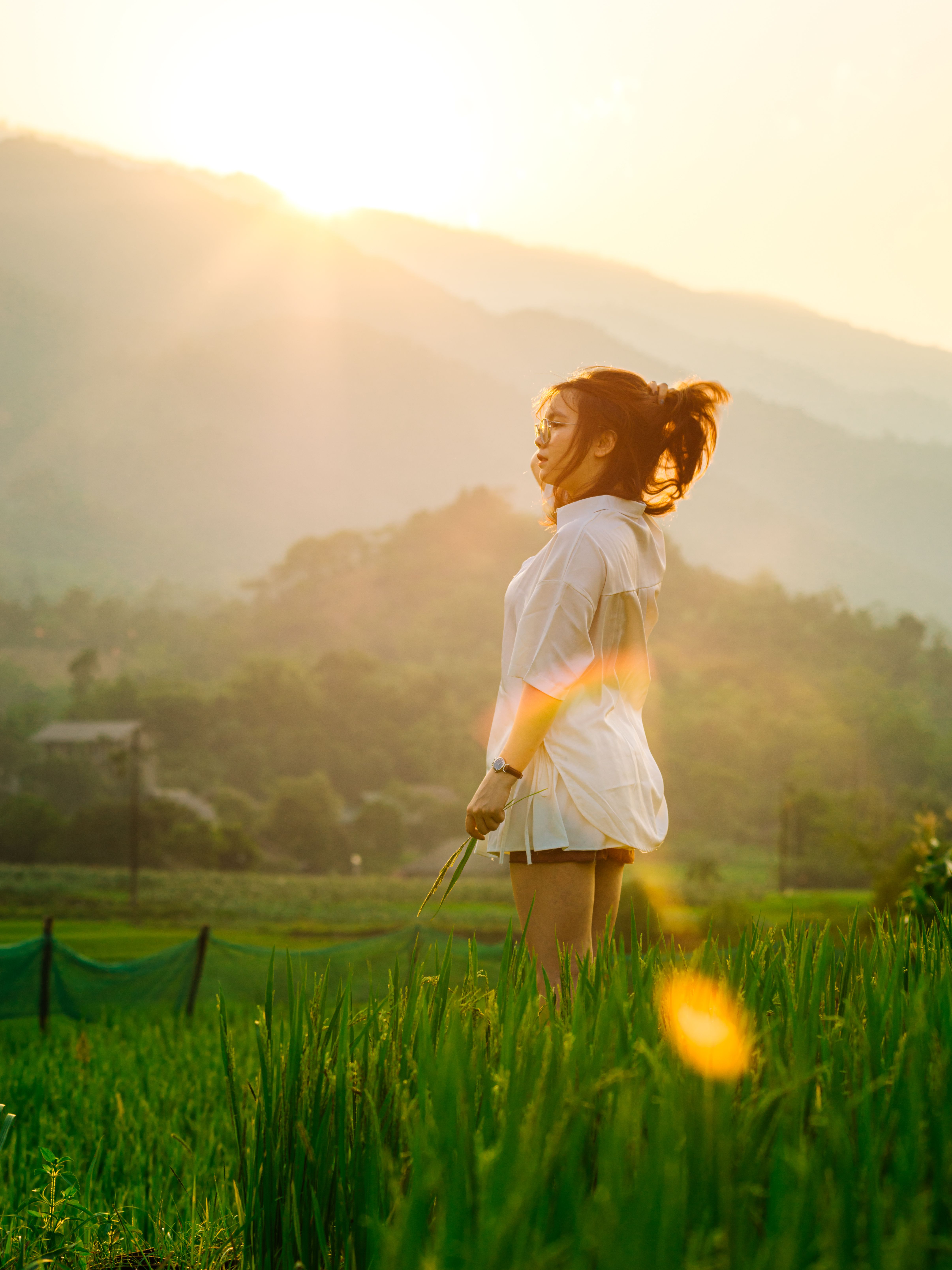 A woman stands in a lush green field at sunset, surrounded by mountains.