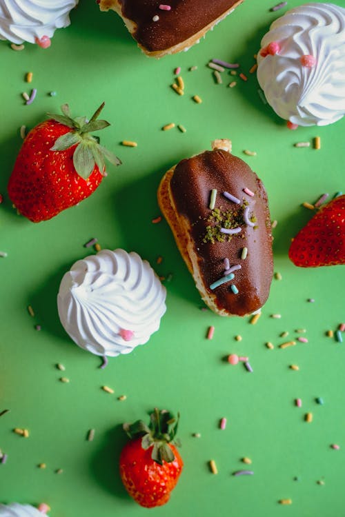 Meringues and Eclairs Topped with Sprinkles Lying in Green Background with Strawberries