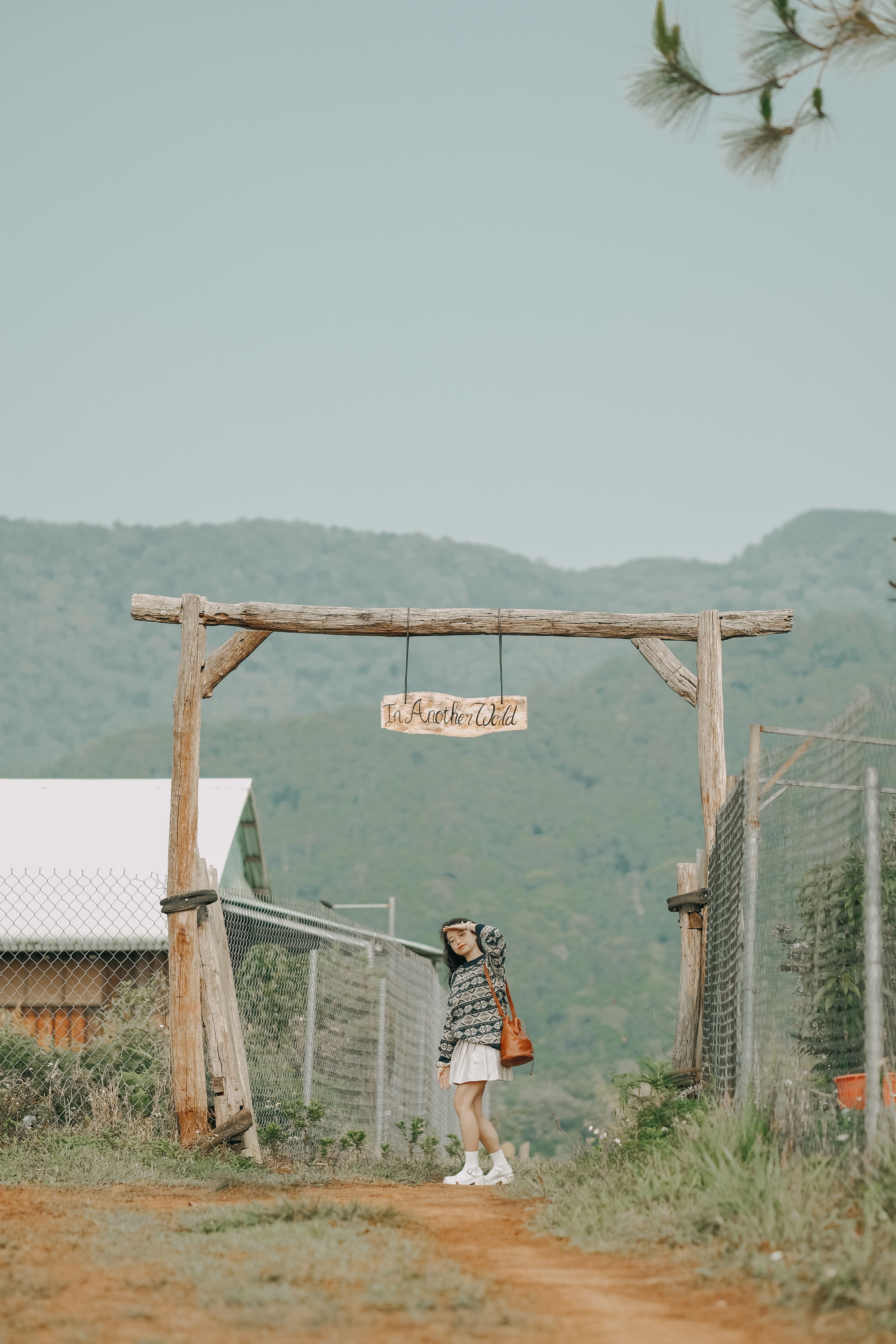 A woman stands beneath a rustic wooden gate in a tranquil rural landscape, embodying peaceful countryside vibes.