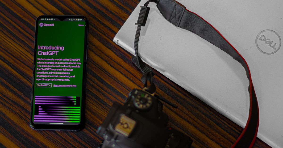 Smartphone with ChatGPT screen next to camera and laptop on wooden desk.