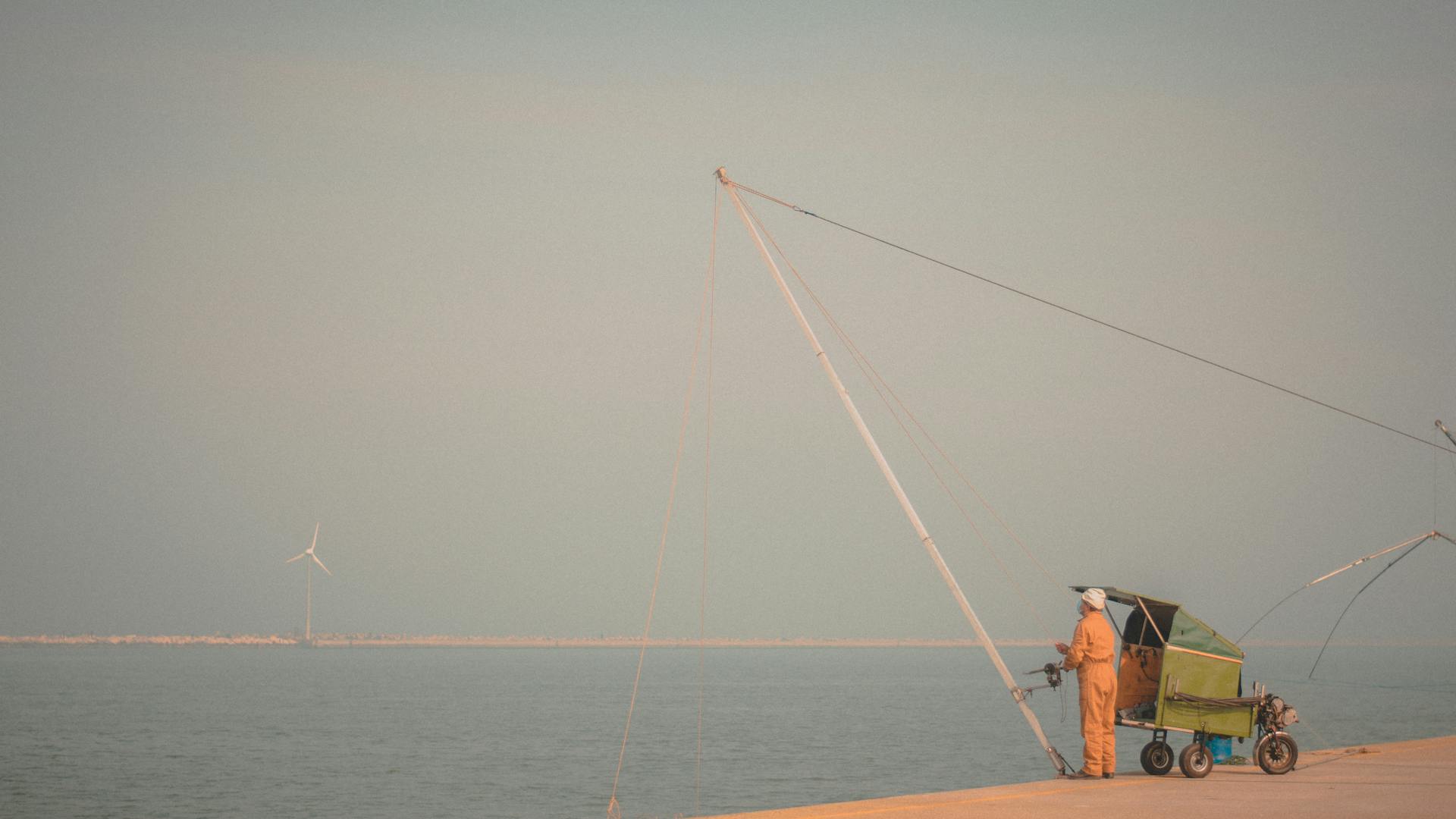 An adult male fishing by the sea with a cart beside him during the day at a harbor.
