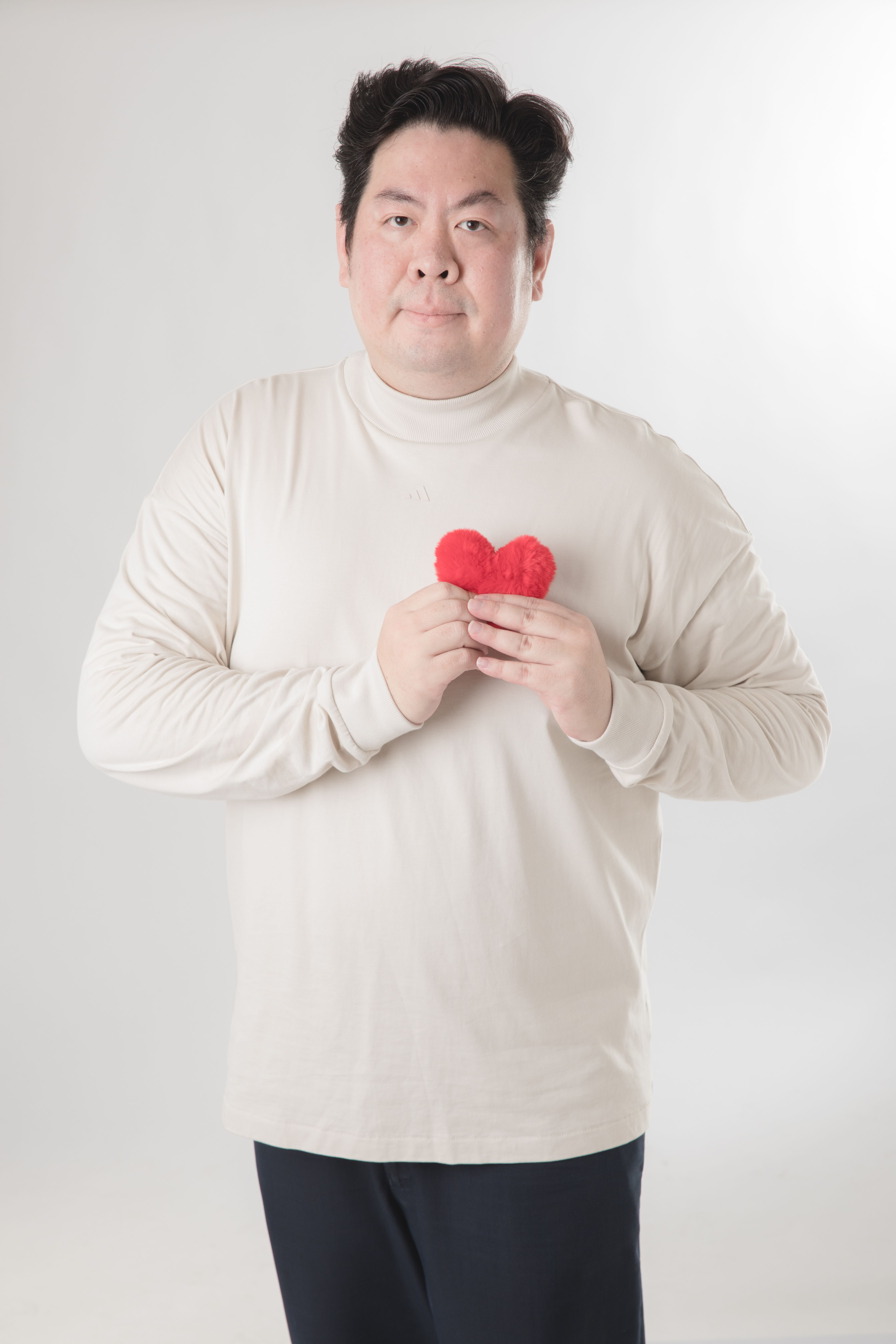 Asian man in a white turtleneck holding a red heart-shaped plush against a white background.