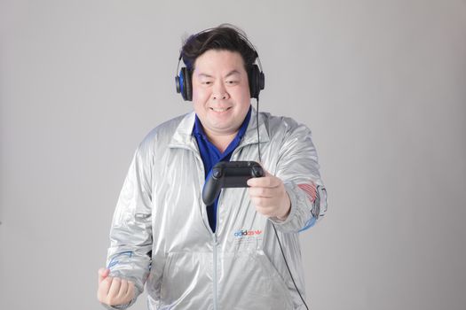 Asian man wearing headphones and silver jacket playing with a controller against a white background.
