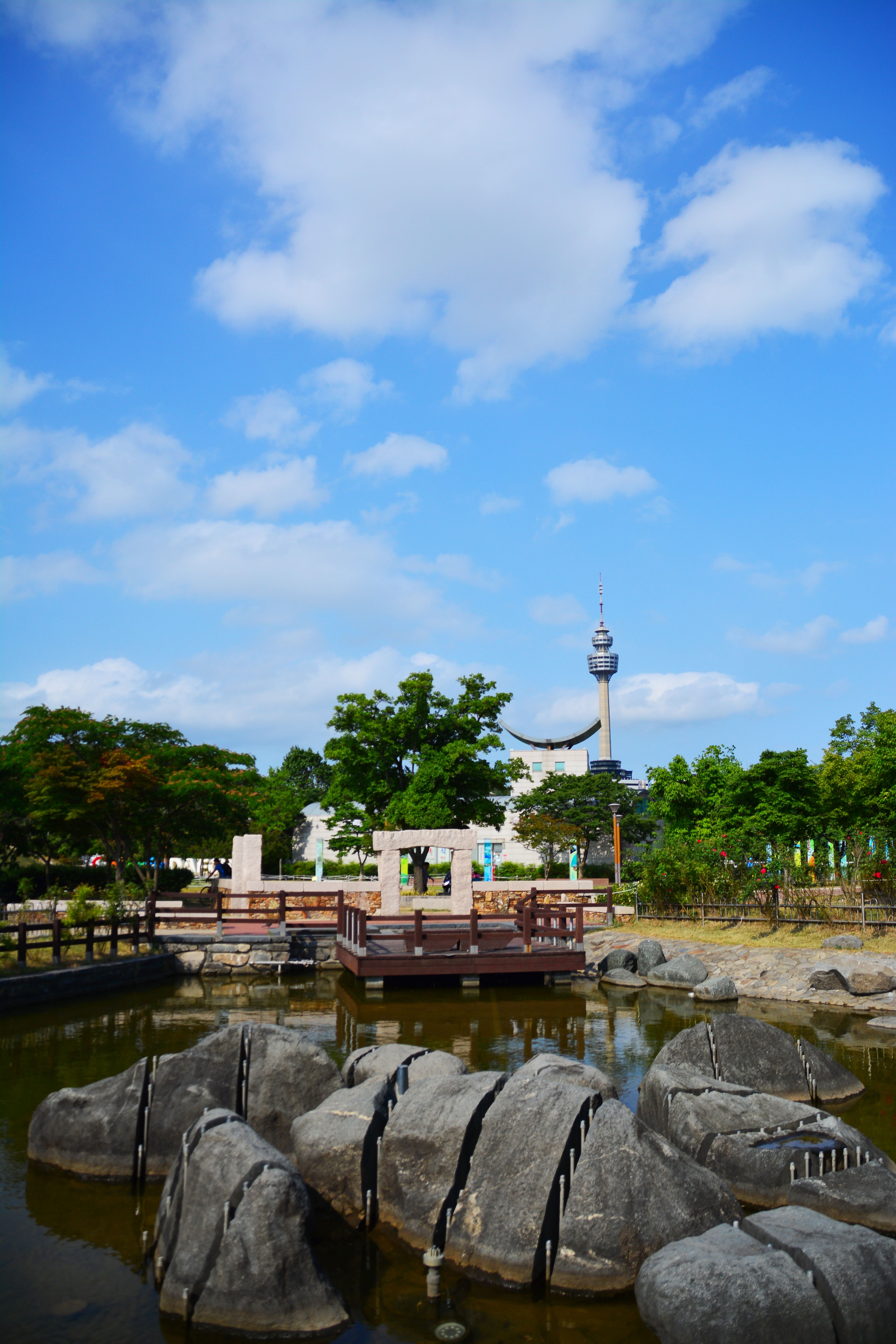 Tranquil park scene with pond, rocks, trees, and a tower under a bright blue sky.