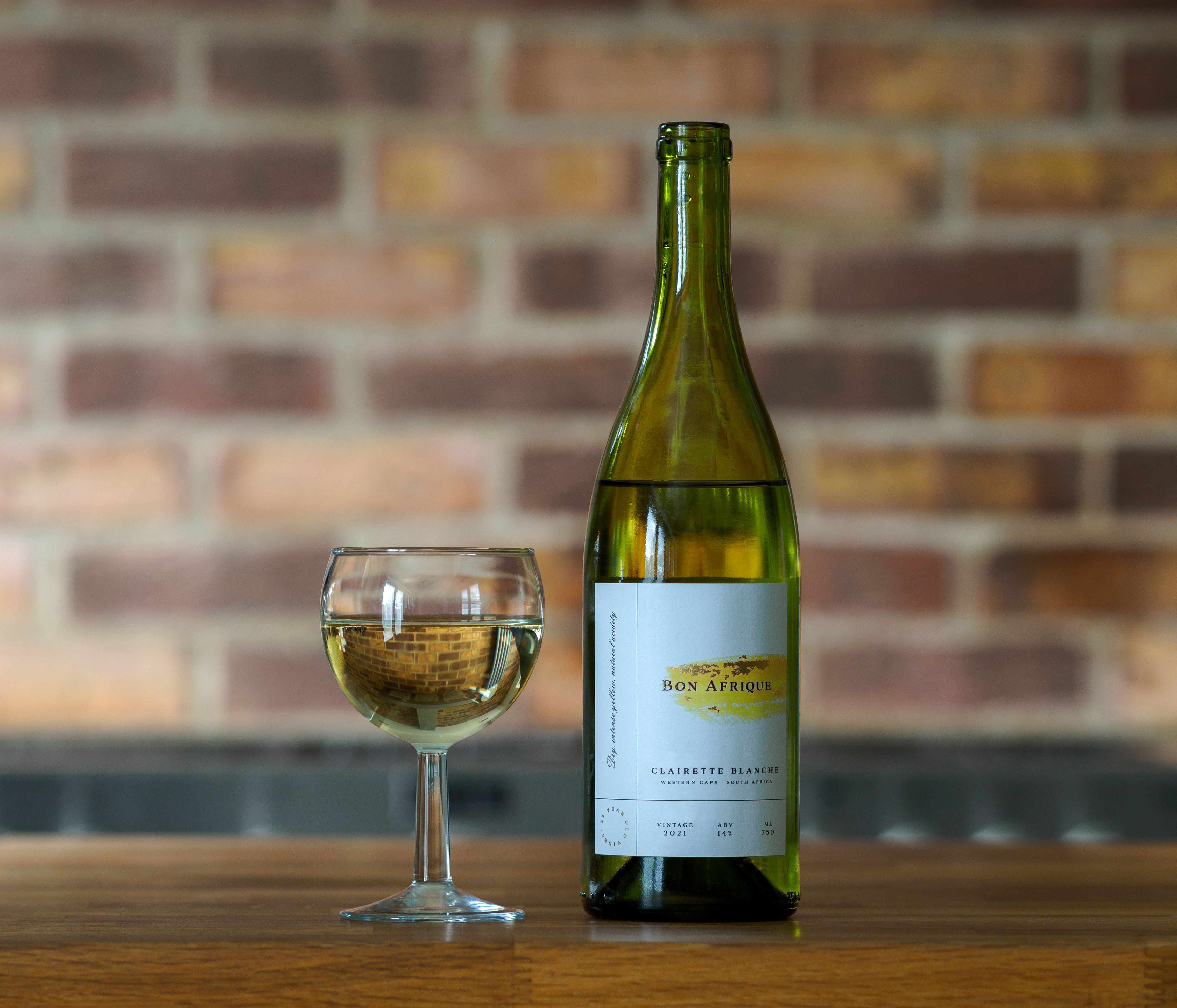 A bottle of white wine and a filled glass on a wooden table against a brick wall.