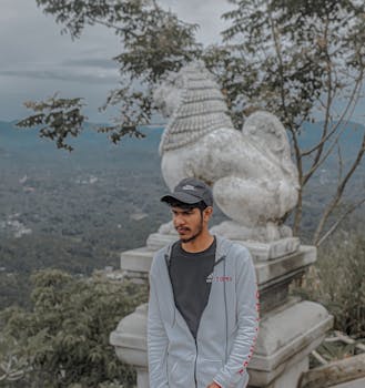 Young man posing by a stone lion statue with a scenic mountain view.