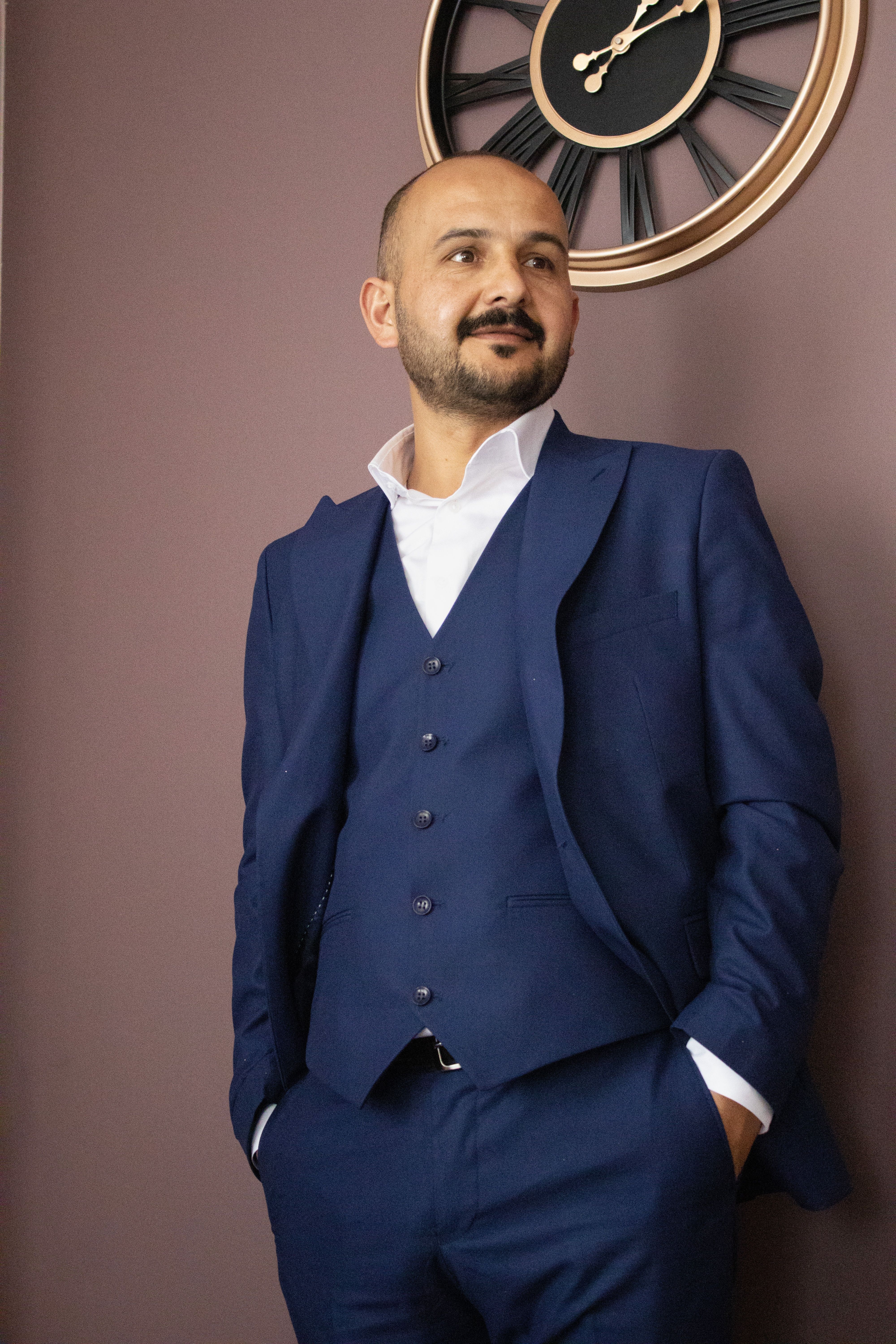 Elegant portrait of a man in a blue suit with a wall clock background, exuding confidence.
