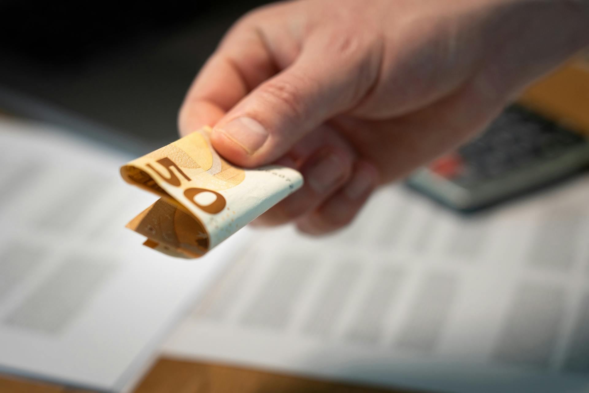 A detailed image of a hand holding a folded 50 euro banknote, with documents in the background.