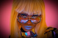 这幅肖像大胆而色彩丰富，描绘了一位戴眼镜、化着妆的时尚女性，背景为粉红色