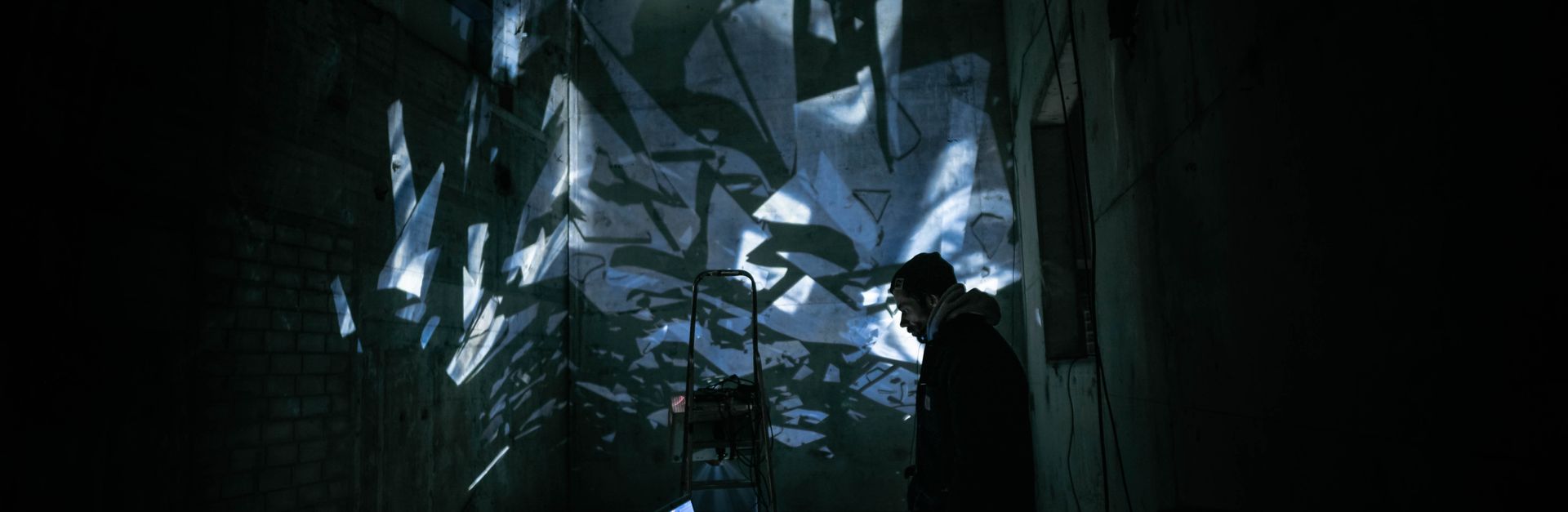 A DJ performing in a dark urban setting with abstract light projections creating a moody atmosphere.