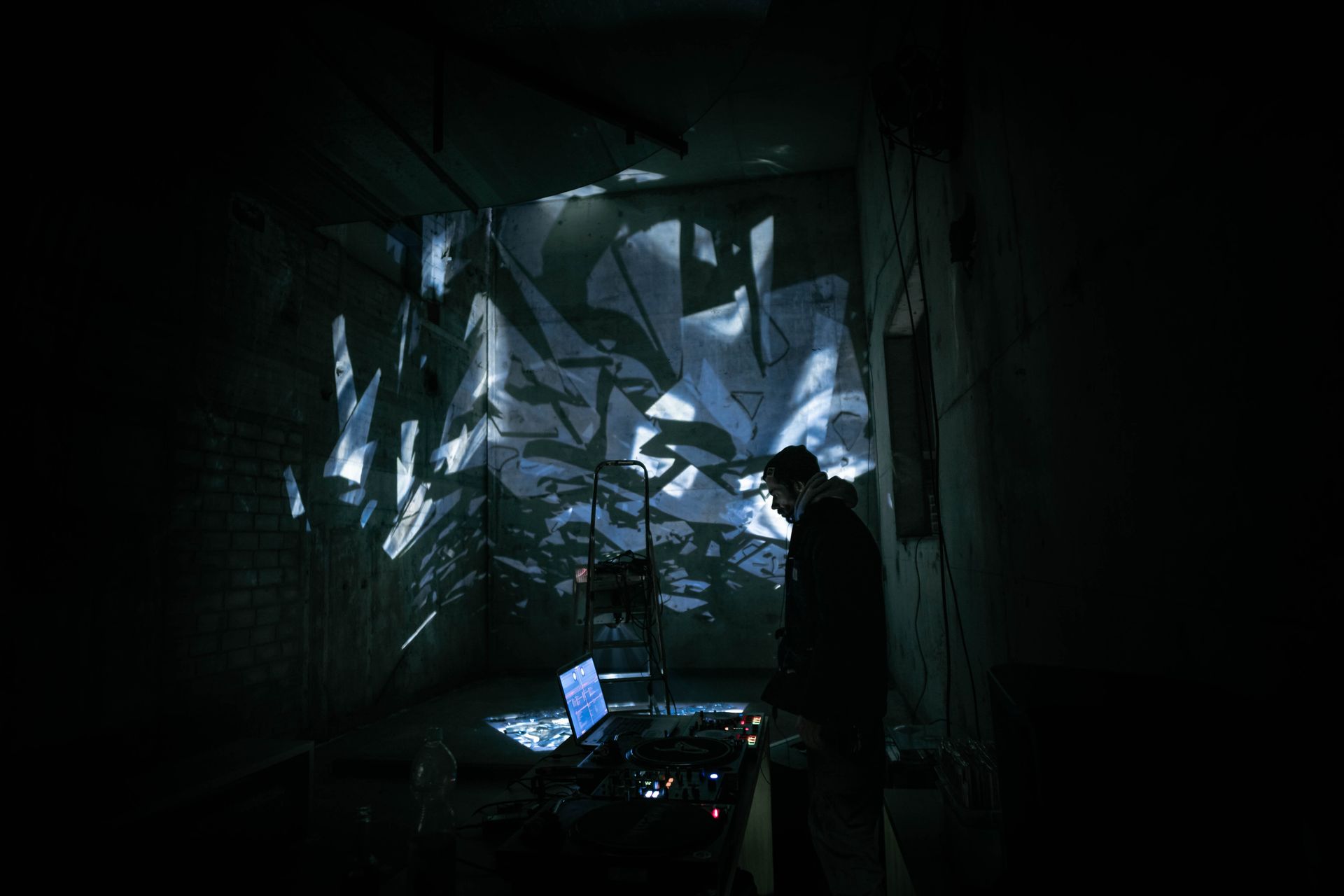 A DJ performing in a dark urban setting with abstract light projections creating a moody atmosphere.