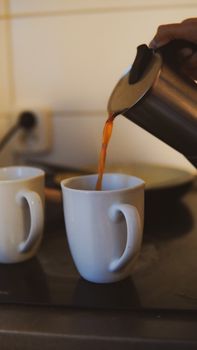 A hand pouring coffee from a metallic pot into a white mug in a kitchen setting.