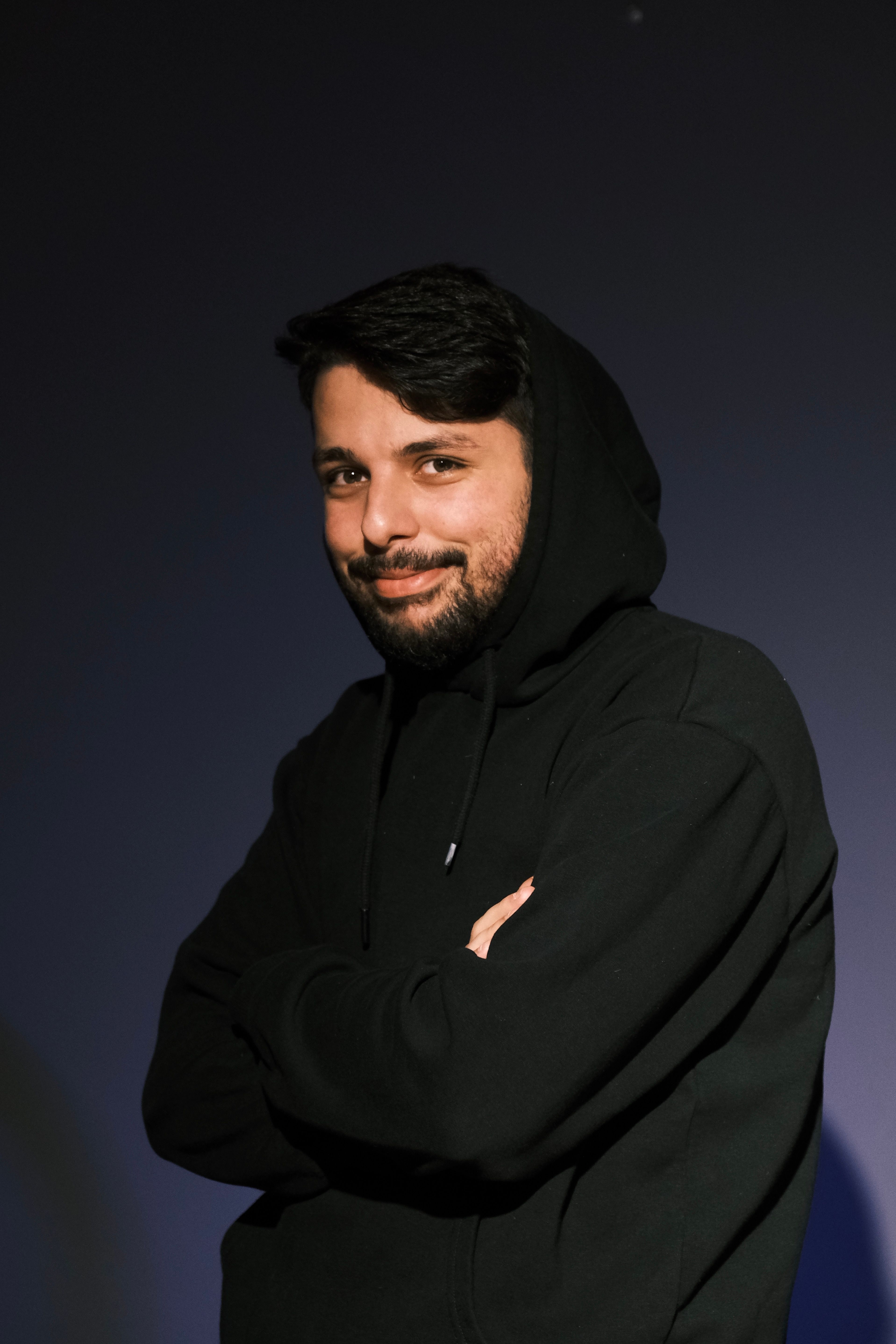A confident man poses in a studio wearing a black hoodie, exuding casual style and charm.
