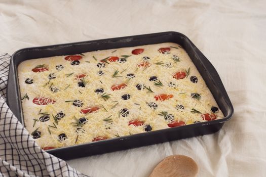 Unbaked focaccia dough topped with cheese, olives, tomatoes, and rosemary.