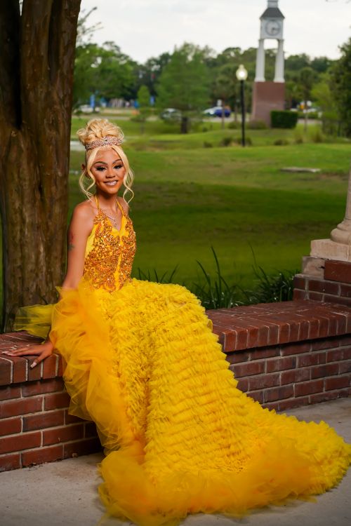 Free A smiling woman in a vibrant yellow dress seated on a brick wall in a park setting. Stock Photo
