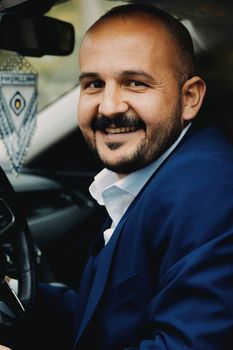 Friendly man with a moustache smiling inside a car, wearing a blue suit.