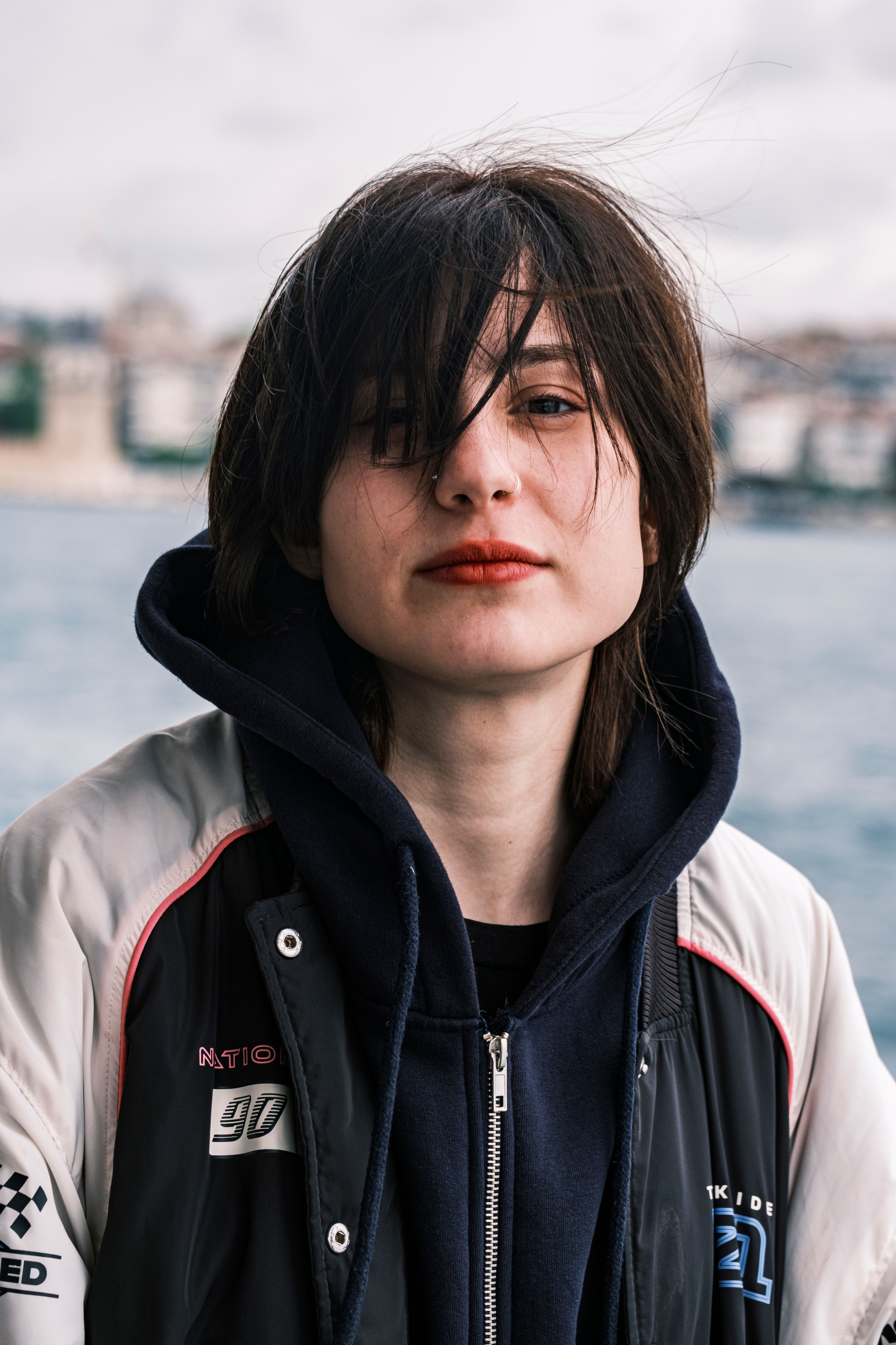 Portrait of a young adult by the shore, wearing a hoodie and jacket on a windy day.