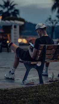A young man casually sits on a bench at dusk using his smartphone in a serene park setting.