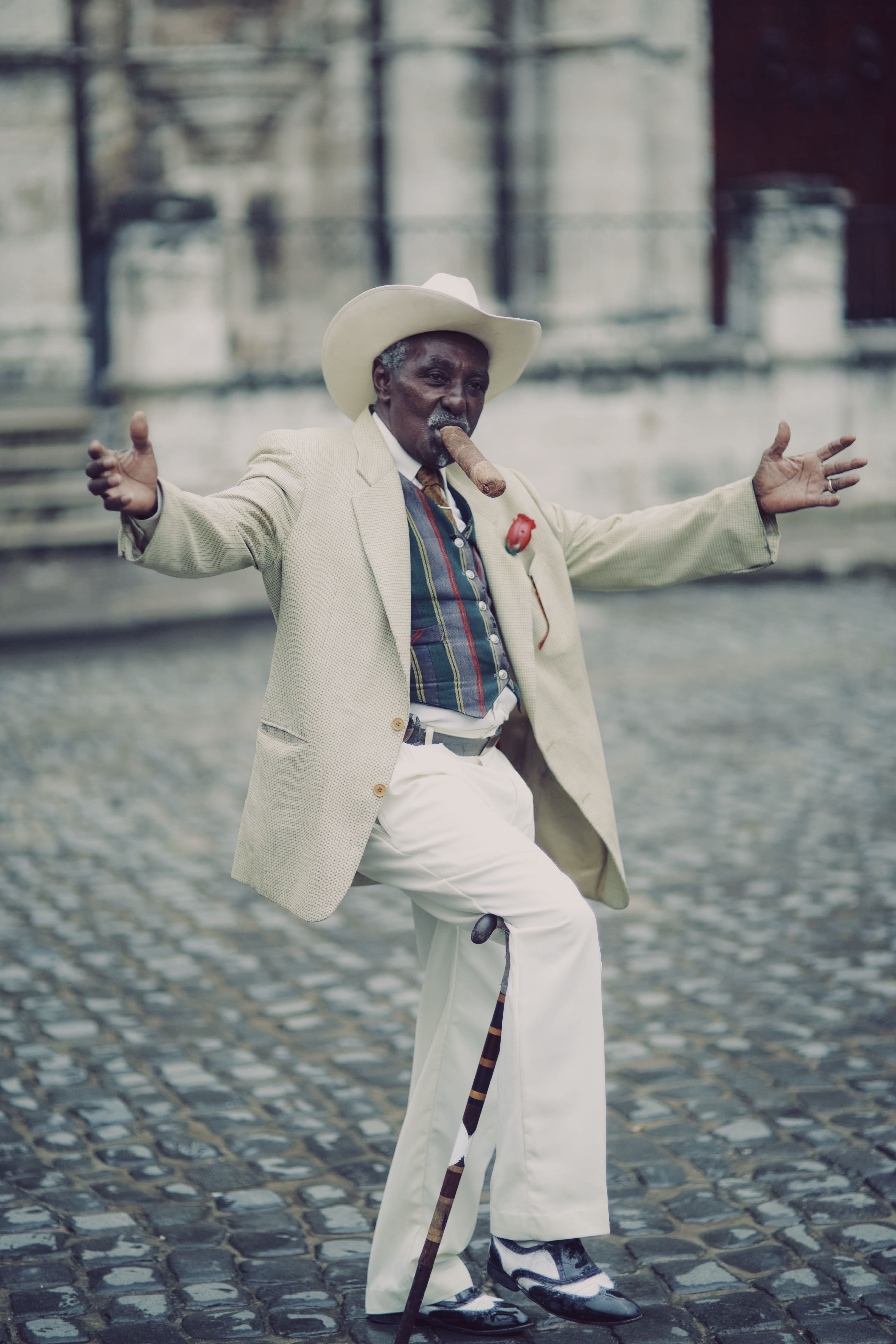 Elderly man dancing joyfully outdoors in elegant suit and hat while smoking a cigar.