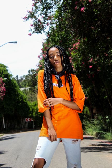 Confident woman in bright orange t-shirt posing on a sunny street with lush greenery.