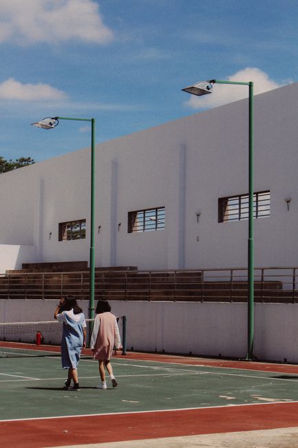 Two women walking on an outdoor tennis court with lamp posts on a sunny day.