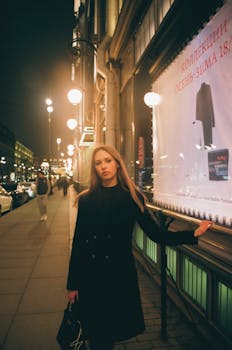 Fashionable woman in a black coat strolling past streetlights in a vibrant city at night.