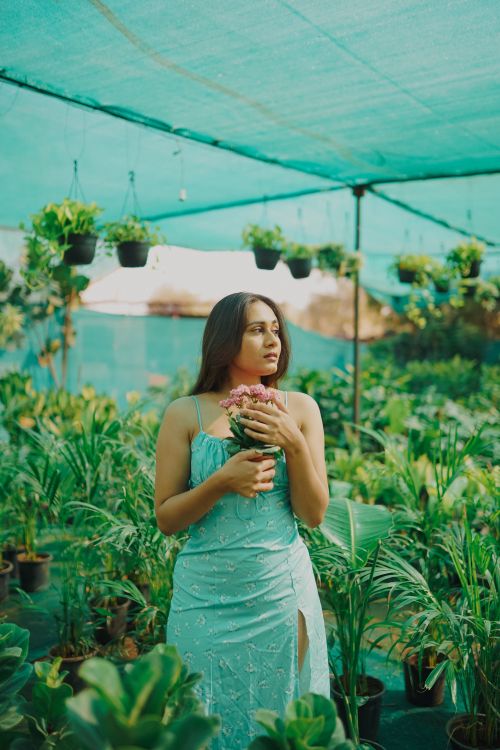 Free Portrait of a woman in a green dress holding flowers in a greenhouse surrounded by plants. Stock Photo