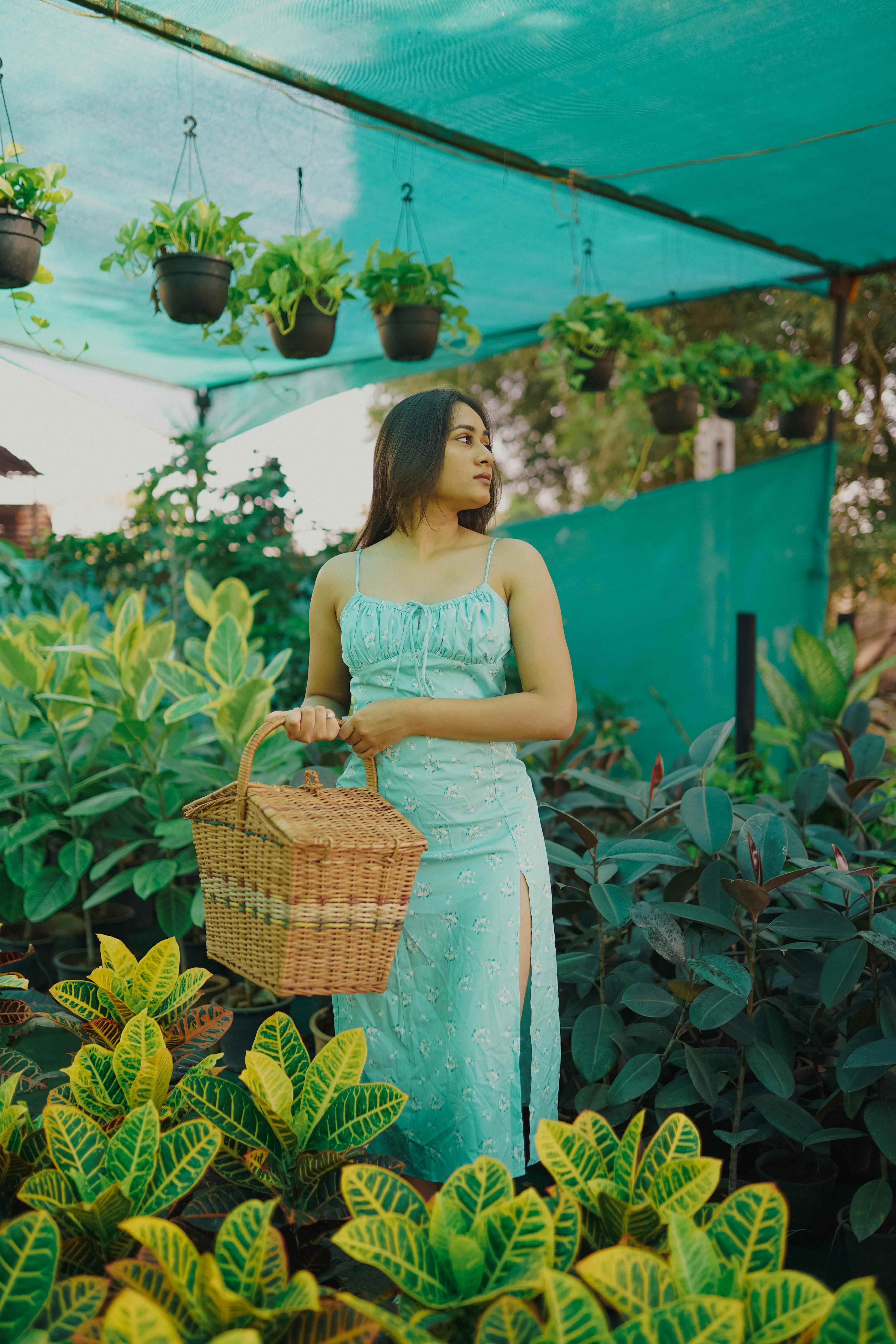 Stylish woman in green dress holding a basket among lush greenhouse plants.