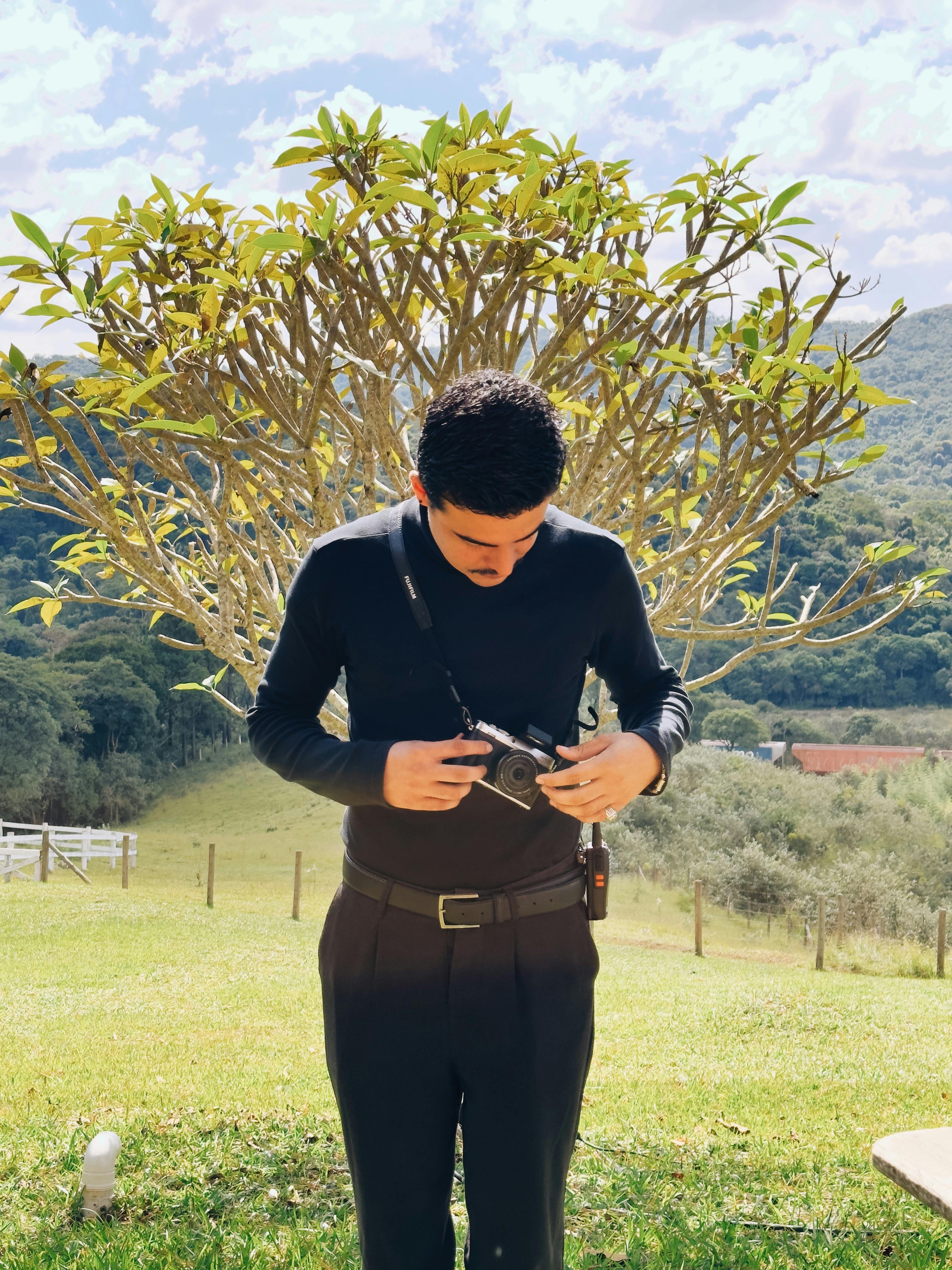 A man stands in a rural setting, examining his vintage camera on a sunny day.