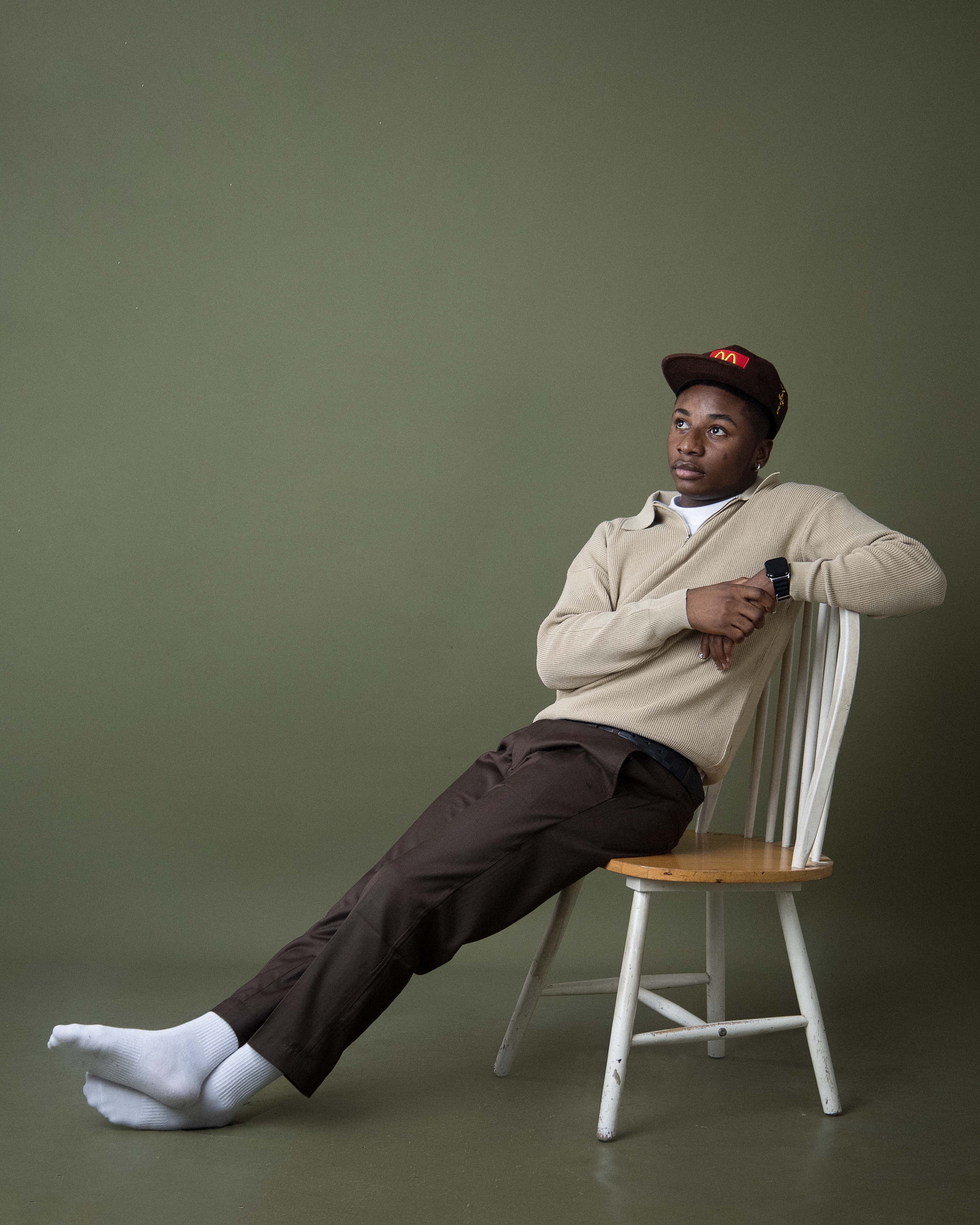 A man in casual attire sitting on a chair against a green background in a studio.