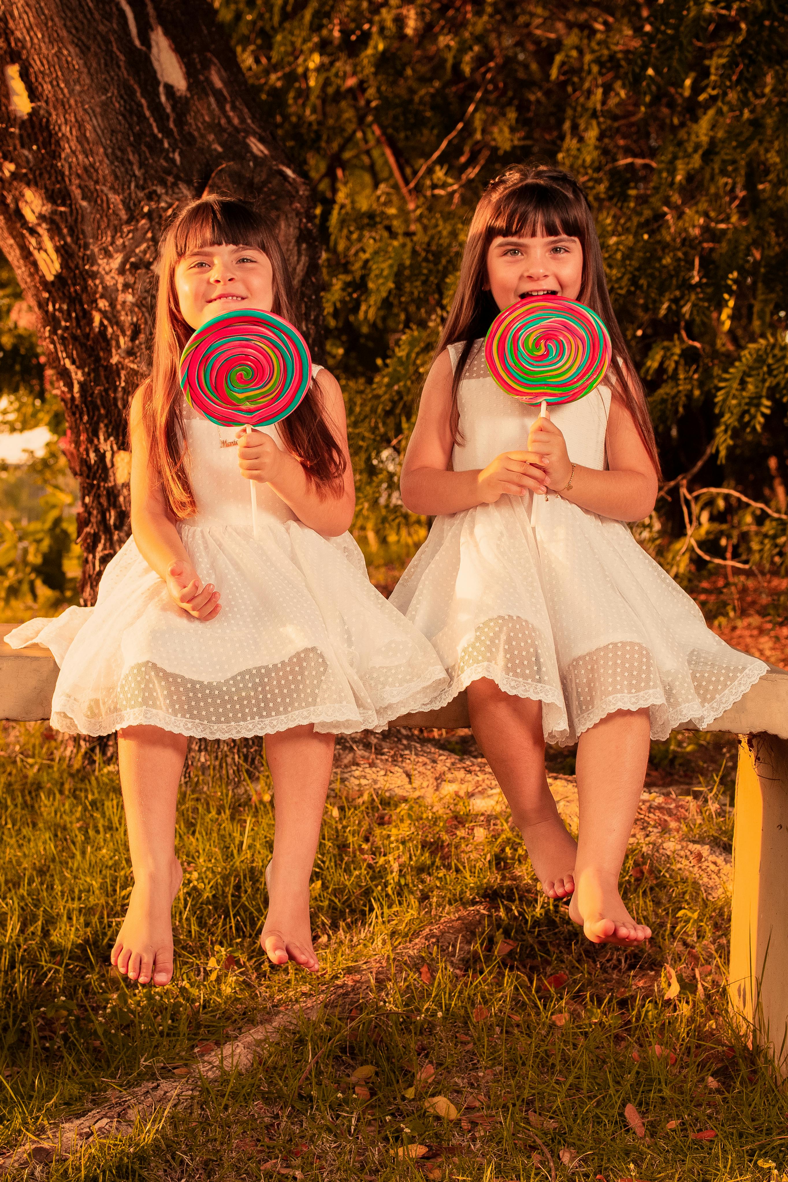 Cute twin girls in white dresses enjoying lollipops, sitting barefoot on a bench in a sunny park.