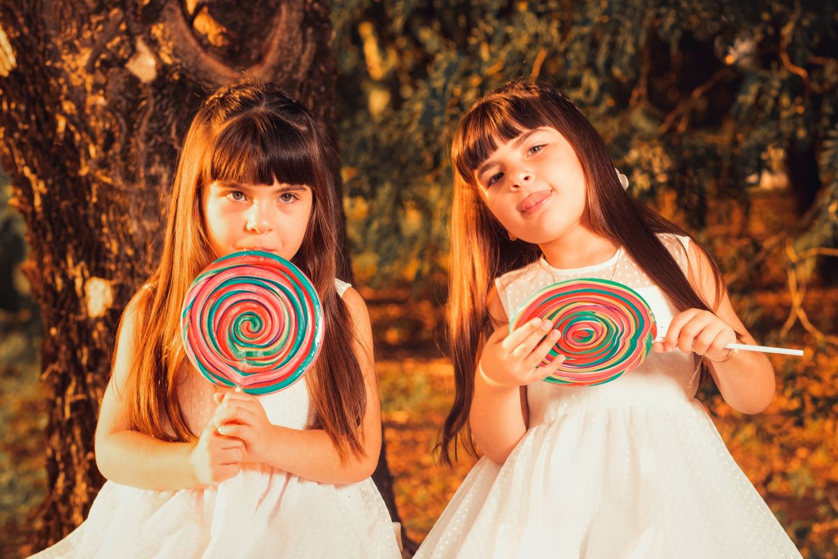 Charming twin girls with brown hair and white dresses enjoying colorful lollipops outdoors in the park.