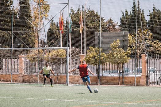 Dynamic soccer match in Málaga, Spain showing player kicking towards goal on sunny day.