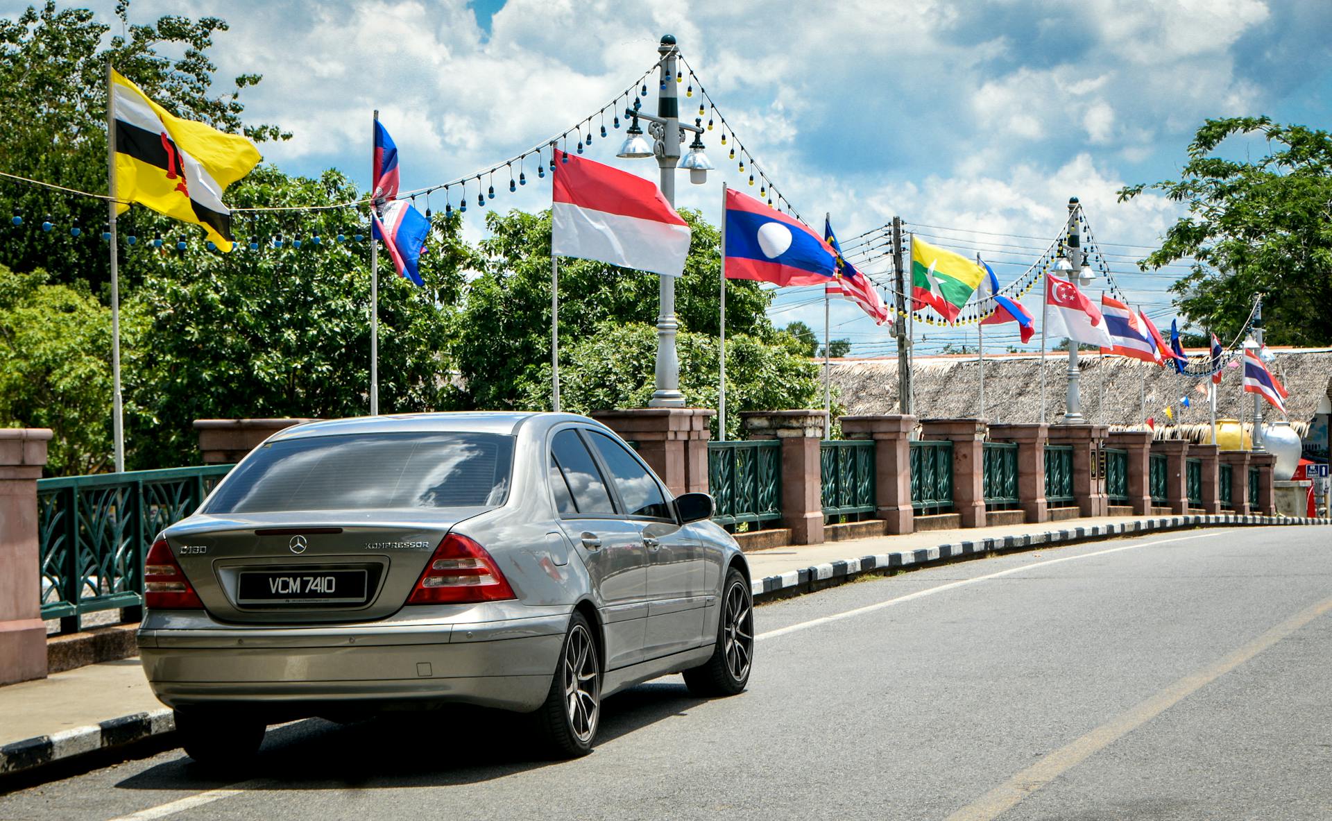 A sleek silver car on a bridge adorned with international flags under a clear blue sky.