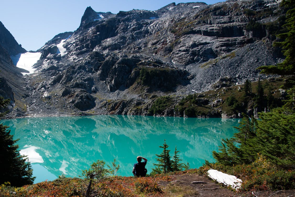 Man Sitting Near Lake Surrounded by Black Rock Formation