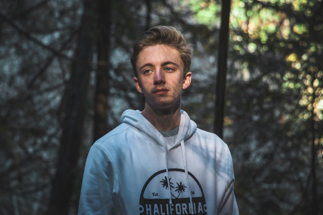 portrait of a young man in a hoodie standing in a sunlit forest showca